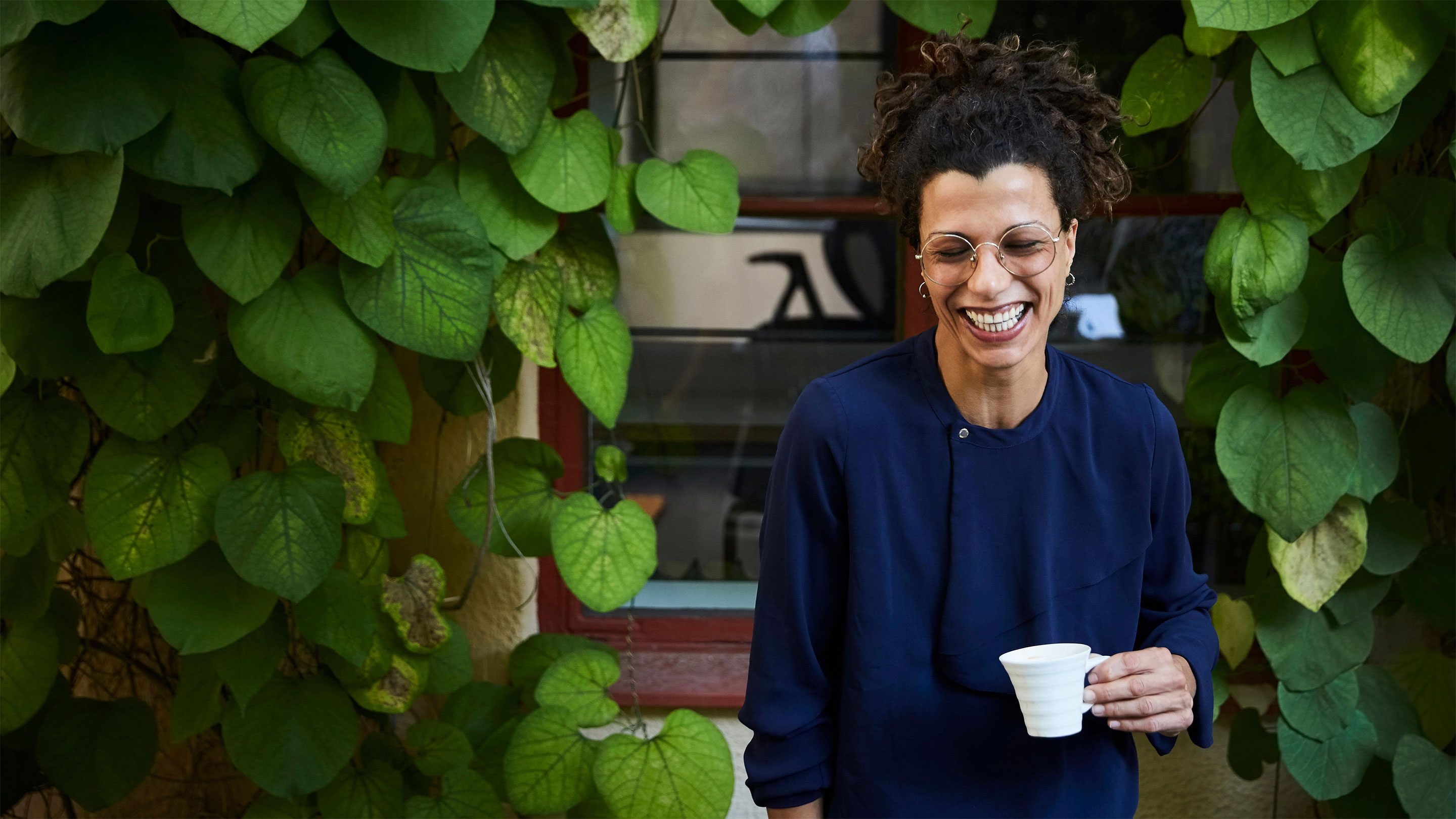 Laughing woman drinking a coffee standing outside of a building covered with foliage 