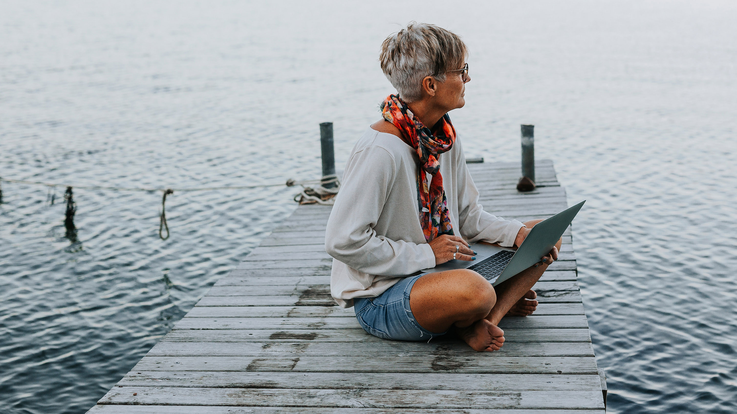 Woman sitting by a lake on a jetty whilst working on her laptop  